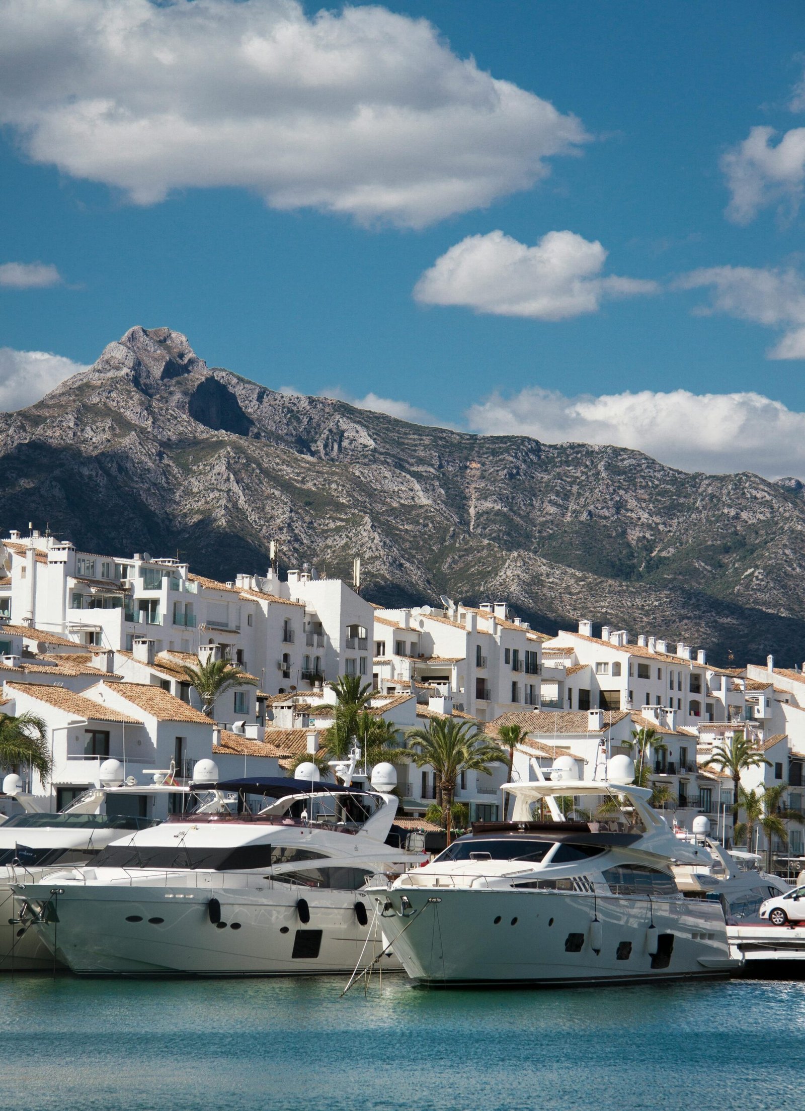 Elegant yachts docked at a Mediterranean harbor with picturesque mountain and town backdrop.